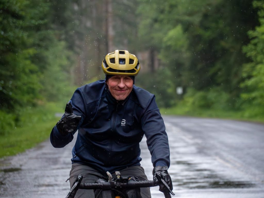 In dark rain gear and a yellow helmet, a middle-aged man smiled and gives the hang loose symbol.