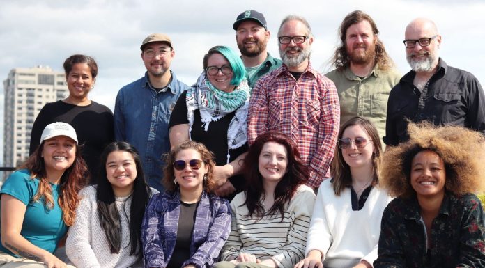 Apply to Join The Urbanist Election Committee The group stands on a rooftop with the Seattle skyline and Elliott Bay in the background.
