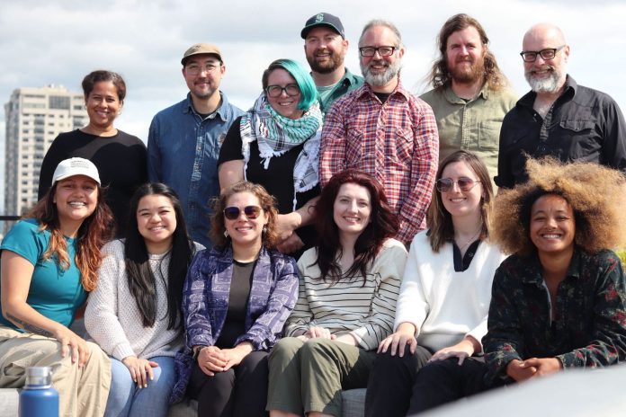 The group stands on a rooftop with the Seattle skyline and Elliott Bay in the background.