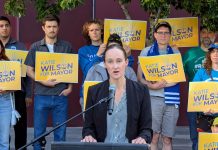 Poll Shows Harrell Is Vulnerable, Wilson Rolls Out Labor Support Wilson stands at a lectern speaking and a dozen supporters stand behind her holding yellow Wilson for Mayor signs on the City Hall Plaza.