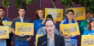 Wilson Defeats Harrell Wilson stands at a lectern speaking and a dozen supporters stand behind her holding yellow Wilson for Mayor signs on the City Hall Plaza.