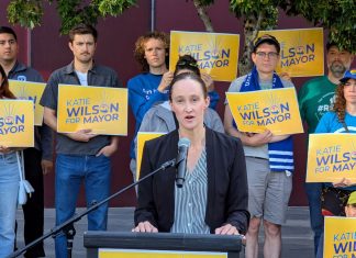 Wilson Defeats Harrell Wilson stands at a lectern speaking and a dozen supporters stand behind her holding yellow Wilson for Mayor signs on the City Hall Plaza.