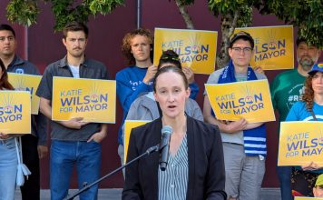 Wilson Defeats Harrell Wilson stands at a lectern speaking and a dozen supporters stand behind her holding yellow Wilson for Mayor signs on the City Hall Plaza.