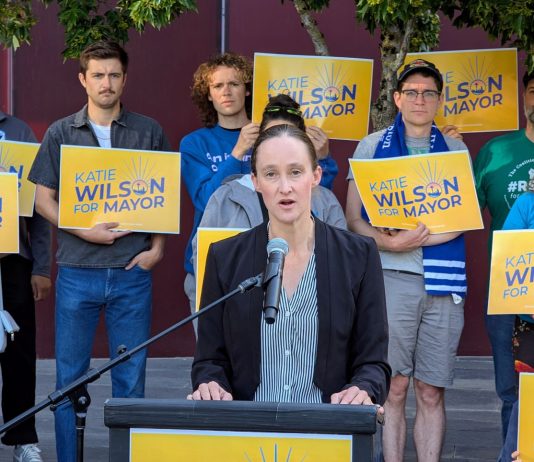 Poll Shows Harrell Is Vulnerable, Wilson Rolls Out Labor Support Wilson stands at a lectern speaking and a dozen supporters stand behind her holding yellow Wilson for Mayor signs on the City Hall Plaza.