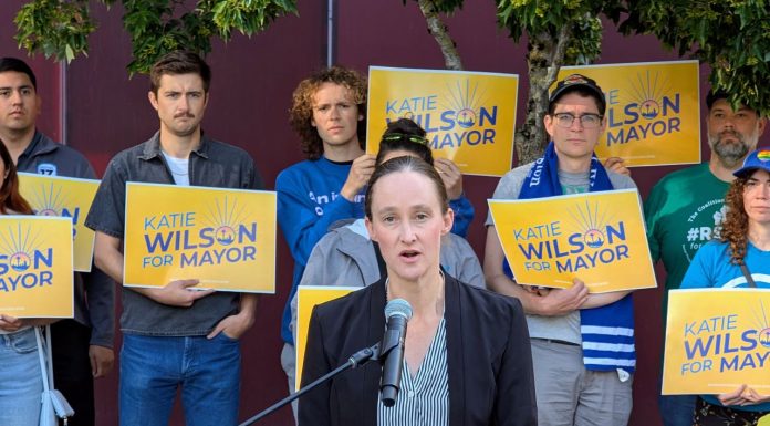 Wilson Defeats Harrell Wilson stands at a lectern speaking and a dozen supporters stand behind her holding yellow Wilson for Mayor signs on the City Hall Plaza.