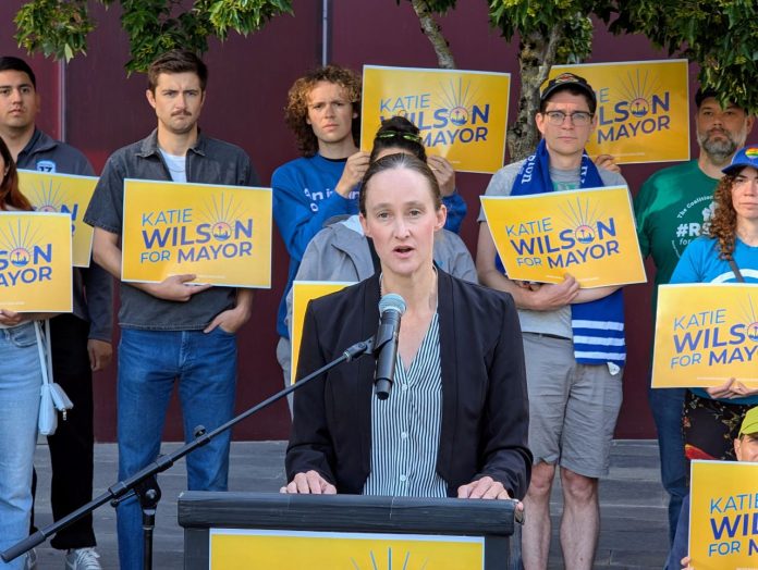 Katie Wilson at City Hall steps union presser - Trumm 20250717 Wilson stands at a lectern speaking and a dozen supporters stand behind her holding yellow Wilson for Mayor signs on the City Hall Plaza.