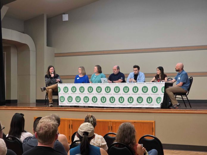 The six panelists sit at a table with the Urbanist U logo on it and Ryna Packer is off to the side moderating.