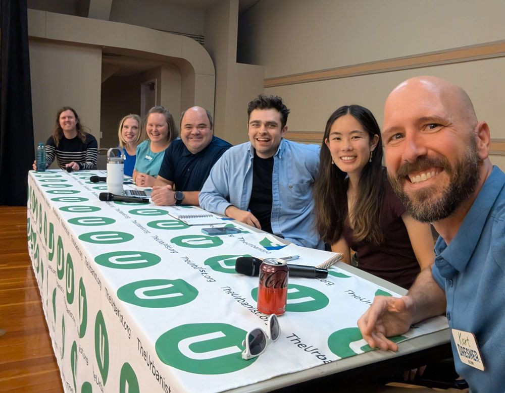 A group of six smiling panelists and one host sit behind a long table covered with a tablecloth that says ‘TheUrbanist.org.’ Kurt is on the far right, holding the camera for a selfie. Microphones, notebooks, and water bottles are on the table, and everyone looks cheerful and engaged.