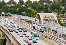 Urbanist Podcast: Crosslake Light Rail + ICE-Triggered Constitutional Crisis An aerial view of the I-90 bridge with a row of westbound cars and a group of construction workers following a light rail vehicle along the center tracks during the tow testing.