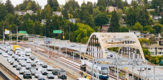 Urbanist Podcast: Crosslake Light Rail + ICE-Triggered Constitutional Crisis An aerial view of the I-90 bridge with a row of westbound cars and a group of construction workers following a light rail vehicle along the center tracks during the tow testing.