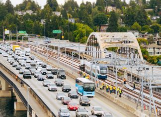 Urbanist Podcast: Crosslake Light Rail + ICE-Triggered Constitutional Crisis An aerial view of the I-90 bridge with a row of westbound cars and a group of construction workers following a light rail vehicle along the center tracks during the tow testing.