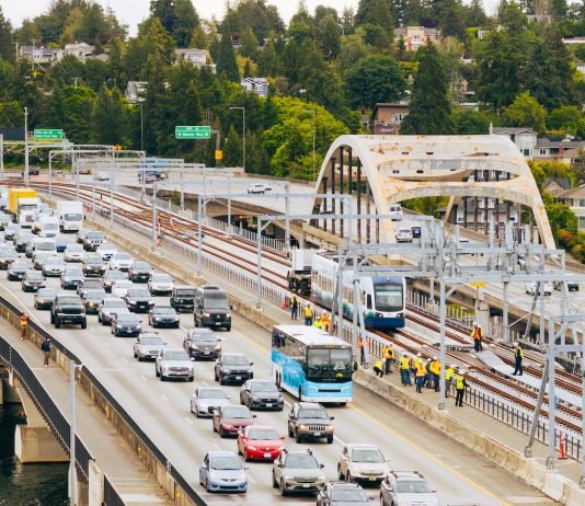 Urbanist Podcast: Crosslake Light Rail + ICE-Triggered Constitutional Crisis An aerial view of the I-90 bridge with a row of westbound cars and a group of construction workers following a light rail vehicle along the center tracks during the tow testing.