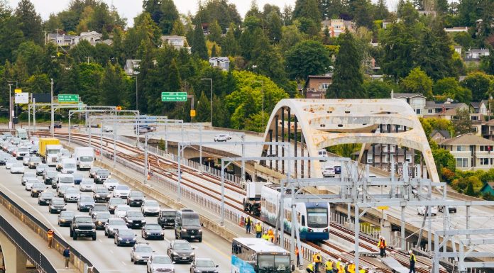 Urbanist Podcast: Crosslake Light Rail + ICE-Triggered Constitutional Crisis An aerial view of the I-90 bridge with a row of westbound cars and a group of construction workers following a light rail vehicle along the center tracks during the tow testing.