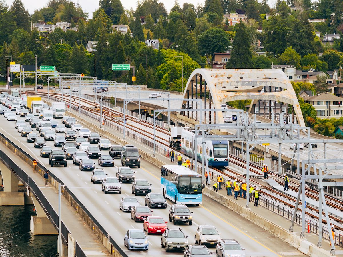 An aerial view of the I-90 bridge with a row of westbound cars and a group of construction workers following a light rail vehicle along the center tracks during the tow testing.