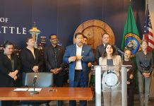Seattle Council Greenlights Rinck’s B&O Tax Overhaul Rinck stands at the lectern and a group of supporters stands behind her, including colleagues Rob Saka and Sara Nelson and Mayor Bruce Harrell.