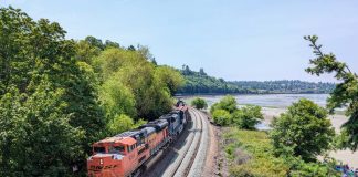 Op-Ed: PCC Provides Model for State Rail Ownership in Washington State A BNSF freight train heads north along the Puget Sound coast, passing under the pedestrian bridge at Carkeek Park. A tree-covered ridge is on the left.