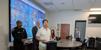 Mayor Bruce Harrell stands at a lectern flanked by Police Chief Shon Barnes and Councilmember Bob Kettle, who chairs the Public Safety Committee. A projection screen displays surveillance footage.