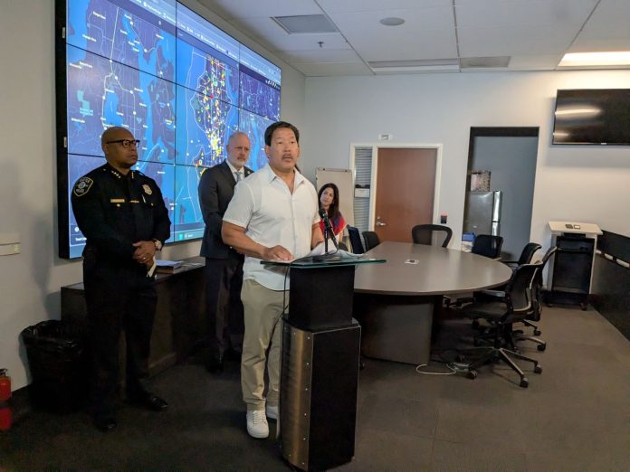 Harrell announces surveillance pilot expansion 20250715 Mayor Bruce Harrell stands at a lectern flanked by Police Chief Shon Barnes and Councilmember Bob Kettle, who chairs the Public Safety Committee. A projection screen displays surveillance footage.