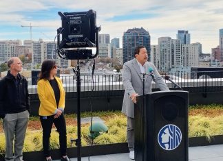 Katie Wilson to Inherit Seattle’s Budget Woes Harrell stands at a lectern with the Seattle City Seal and the skyline visible behind him from a midrise rooftop.