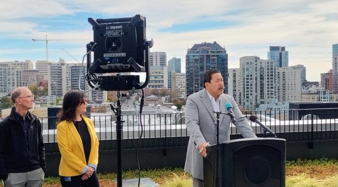 Katie Wilson to Inherit Seattle’s Budget Woes Harrell stands at a lectern with the Seattle City Seal and the skyline visible behind him from a midrise rooftop.