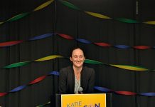Katie Wilson Leads Bruce Harrell in Early Primary Election Results Wilson stands in front of a lectern with her yellow campaign sign with rainbow streamers behind her.