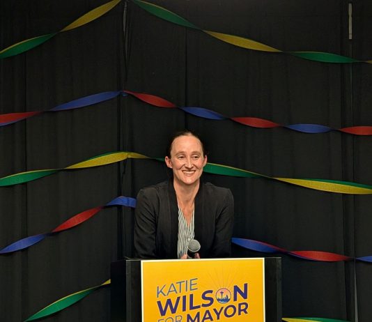 Katie Wilson Leads Bruce Harrell in Early Primary Election Results Wilson stands in front of a lectern with her yellow campaign sign with rainbow streamers behind her.