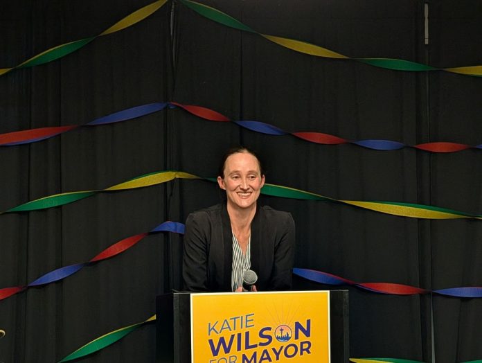 Katie Wilson primary victory speech 1600px Trumm Wilson stands in front of a lectern with her yellow campaign sign with rainbow streamers behind her.