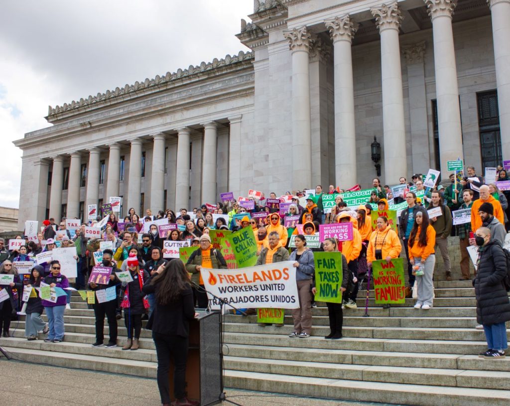 More than 100 advocates hold signs urging a balanced tax code on the steps of the state Capitol building with its many marble columns.