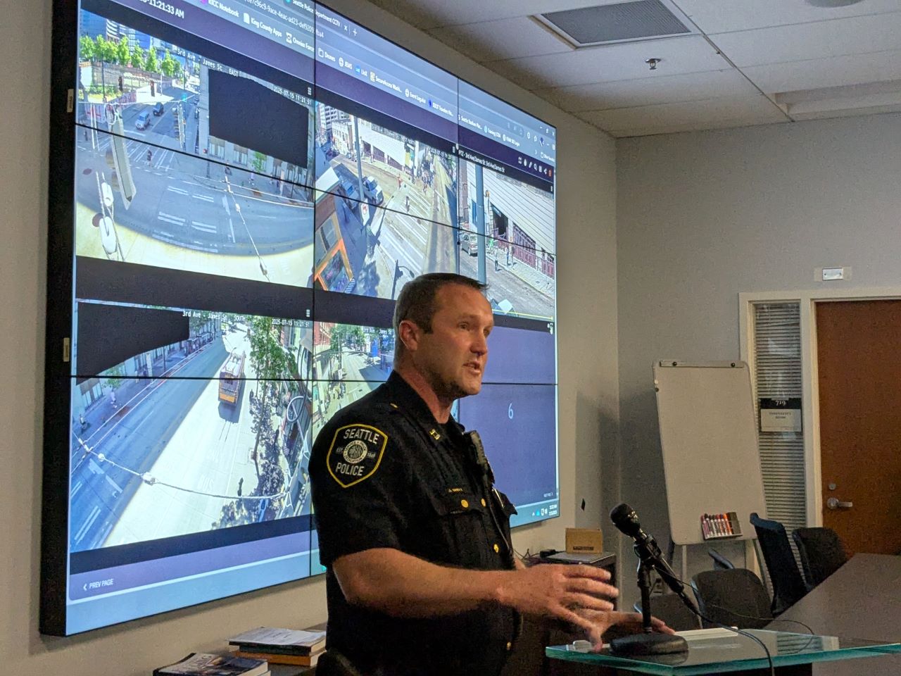 SPD captain Britt wears his police uniform and stands in front of a projection screen playing a feed of surveillance footage.