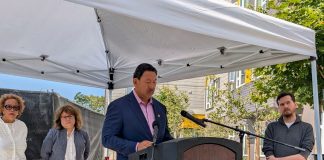 Harrell stands at a lectern under a white tent with Bryant Manor apartment building in the background.