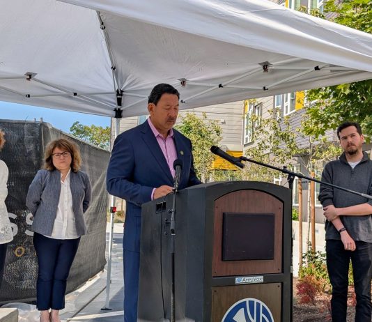 Harrell Announces Reparations Fund, Ramps Up Attacks on Wilson Harrell stands at a lectern under a white tent with Bryant Manor apartment building in the background.