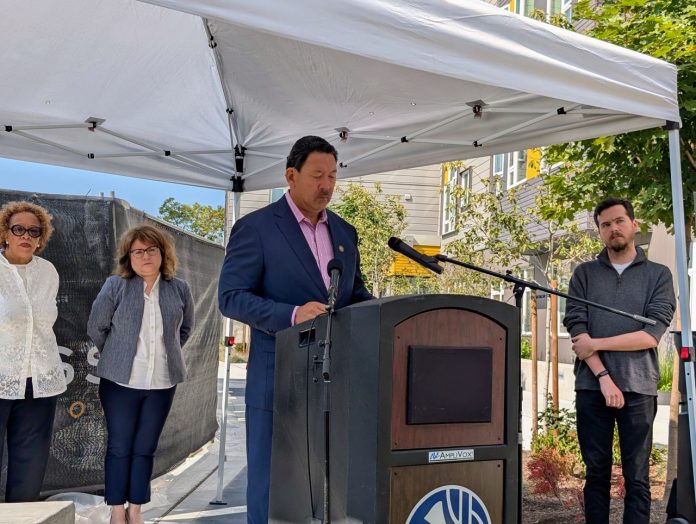 Harrell stands at a lectern under a white tent with Bryant Manor apartment building in the background.