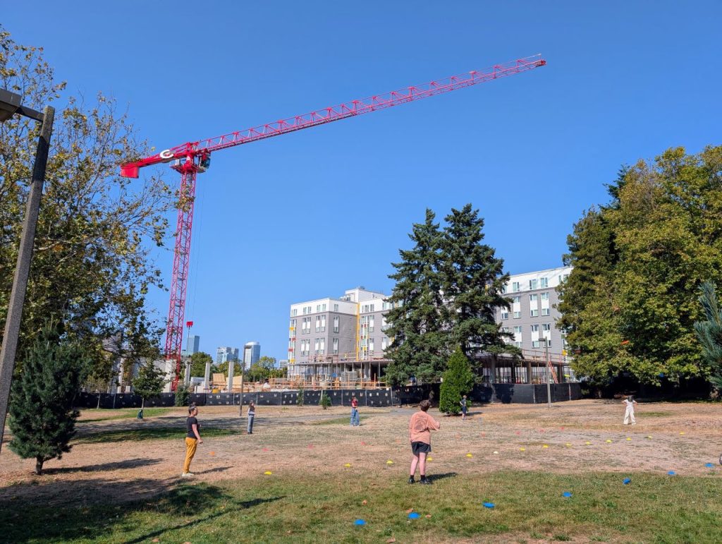 A group of six plays frisbee in the park with the construction crane and skyline looming in the distance.