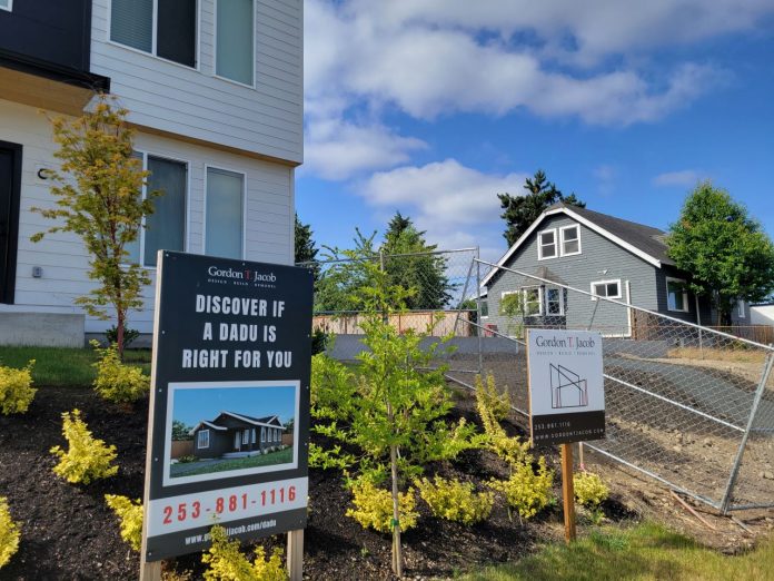A sign says "Discover if an ADU is right for you" in front of a home with a backyard cottage.