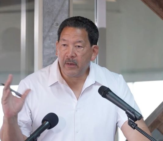 In Last Stand, Harrell Intensifies Negative Attacks on Wilson Wearing a white polo Harrell stands at a lectern gesturing with a pen in his hand.