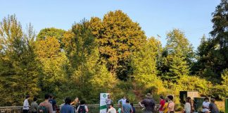 Kicking off The Urbanist Fall Subscriber Drive, Score Your Merch A group of about 30 people stands in a park with an Urbanist banner set up and a stand of trees in the background.