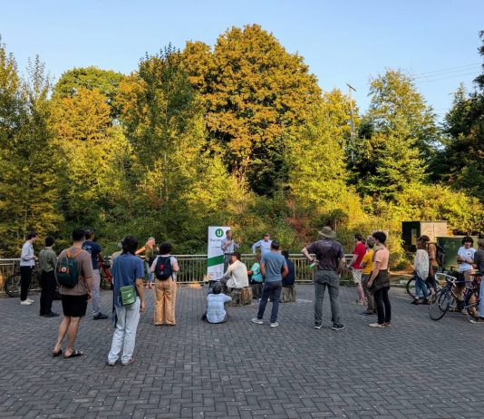 Kicking off The Urbanist Fall Subscriber Drive, Score Your Merch A group of about 30 people stands in a park with an Urbanist banner set up and a stand of trees in the background.