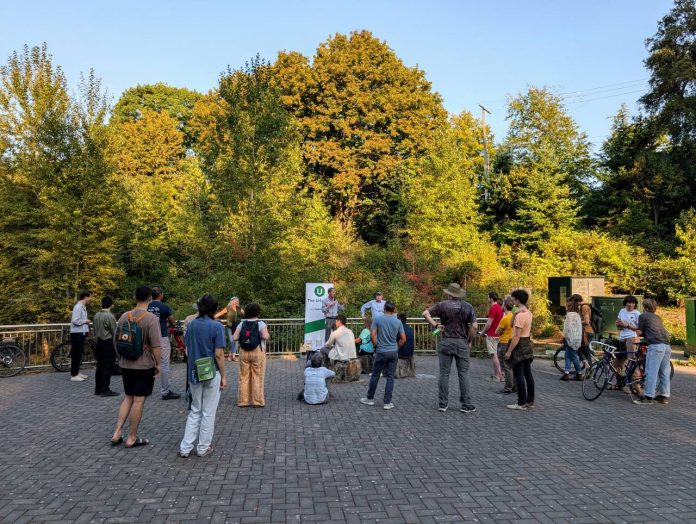 A group of about 30 people stands in a park with an Urbanist banner set up and a stand of trees in the background.