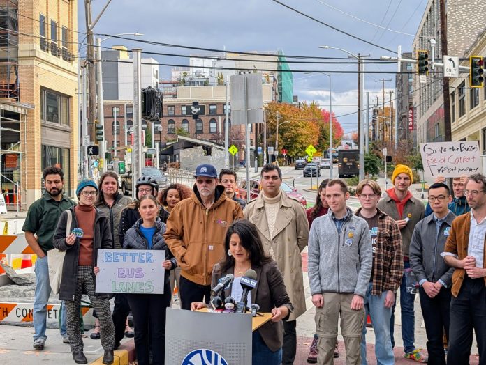 Alexis Rinck announces Better Bus Lanes campaign - Trumm 20251022 More than two dozen advocates stand behind Rinck at the bus stop at 12th Avenue and Union Street, with Madison Street in the background.
