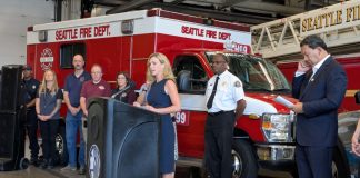 Barden stands at a lectern at a fire hall with a fire truck, fire chief, mayor, and CARE staff in the background.