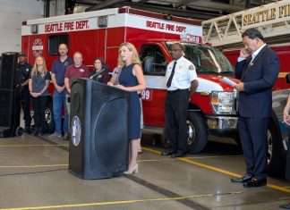 Sales Tax Hike Would Fund Civilian Responder Expansion, Barring SPOG Obstruction Barden stands at a lectern at a fire hall with a fire truck, fire chief, mayor, and CARE staff in the background.