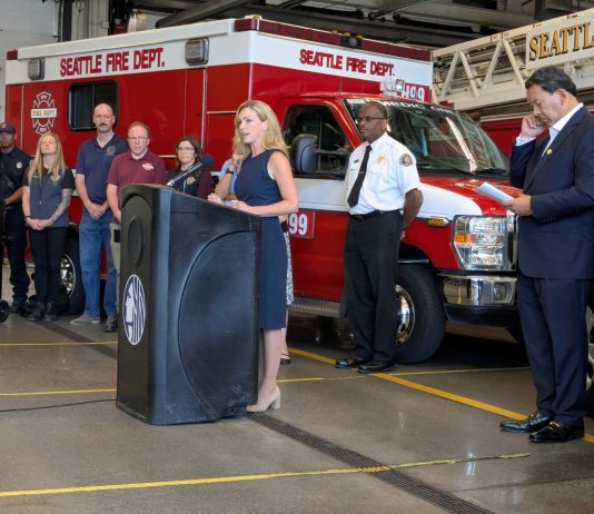 Sales Tax Hike Would Fund Civilian Responder Expansion, Barring SPOG Obstruction Barden stands at a lectern at a fire hall with a fire truck, fire chief, mayor, and CARE staff in the background.