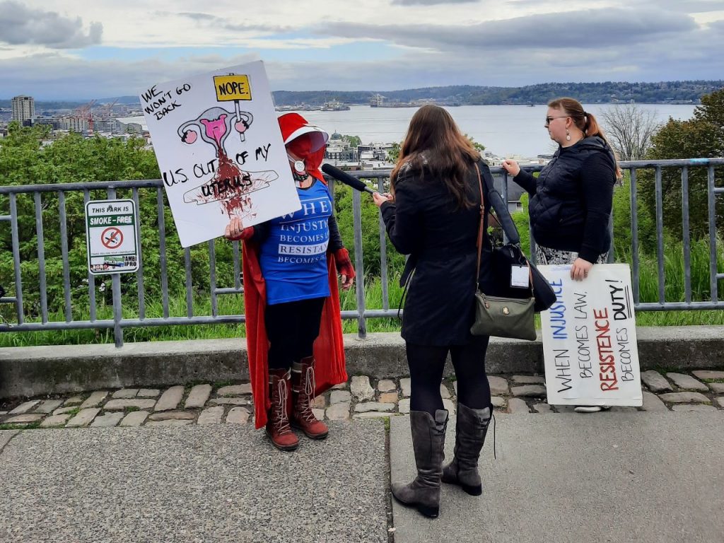 A woman wearing a bright red cape in the style of Handmaids Tale holds a sign showing a cartoon uterus and saying "We won't go back. Nope. U.S. out my uterus." She is being interviewed by a reporter at Kerry Park.