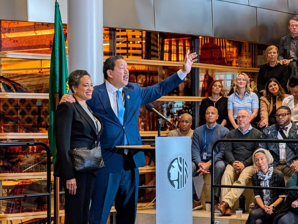 Harrell waves goodbye at the end of his speech with his wife on his arm. They stand at a lectern in the lobby of city hall.