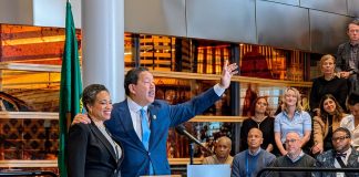 Harrell Concedes to Wilson, Ushering in a New Era in Seattle Harrell waves goodbye at the end of his speech with his wife on his arm. They stand at a lectern in the lobby of city hall.