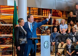 Harrell Concedes to Wilson, Ushering in a New Era in Seattle Harrell waves goodbye at the end of his speech with his wife on his arm. They stand at a lectern in the lobby of city hall.