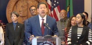 Harrell Vows to Block ICE Overreach, but Plan Lacks Teeth Harrell wears a suit and stands at a lectern with the seal of Seattle. He's surrounded by city councilmembers and staffers including Sara Nelson, Rob Saka, and Mark Solomon.