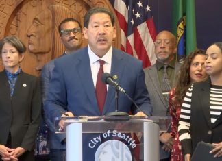 Harrell Vows to Block ICE Overreach, but Plan Lacks Teeth Harrell wears a suit and stands at a lectern with the seal of Seattle. He's surrounded by city councilmembers and staffers including Sara Nelson, Rob Saka, and Mark Solomon.