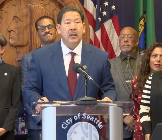 Harrell Vows to Block ICE Overreach, but Plan Lacks Teeth Harrell wears a suit and stands at a lectern with the seal of Seattle. He's surrounded by city councilmembers and staffers including Sara Nelson, Rob Saka, and Mark Solomon.