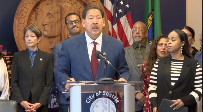 Harrell Vows to Block ICE Overreach, but Plan Lacks Teeth Harrell wears a suit and stands at a lectern with the seal of Seattle. He's surrounded by city councilmembers and staffers including Sara Nelson, Rob Saka, and Mark Solomon.